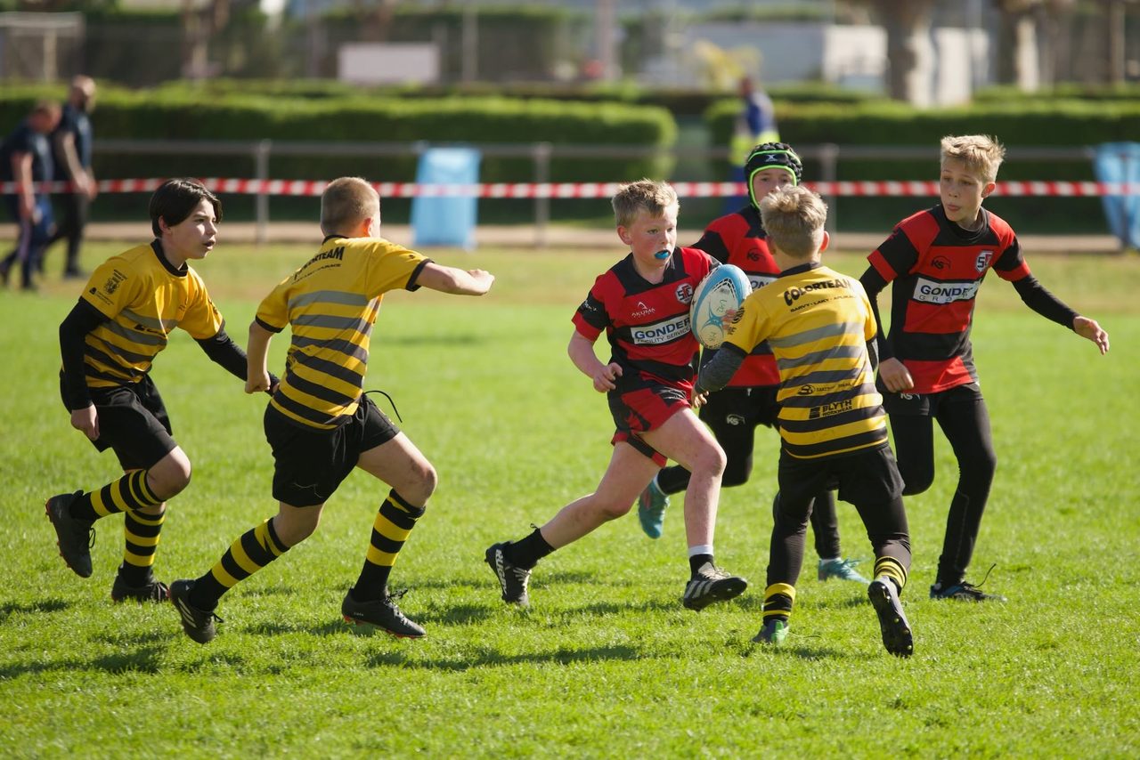 Two teams in red and black and in yellow and black stripes contest the ball in a youth match on the grass pitch